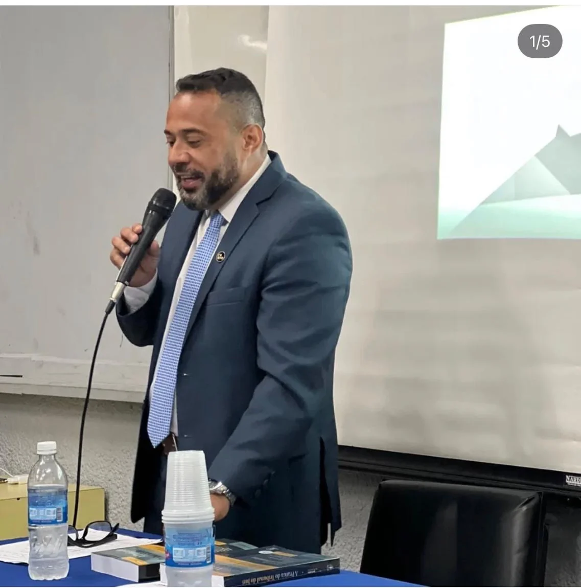 Alexandre Domingues em palestra, de traje social, ao microfone